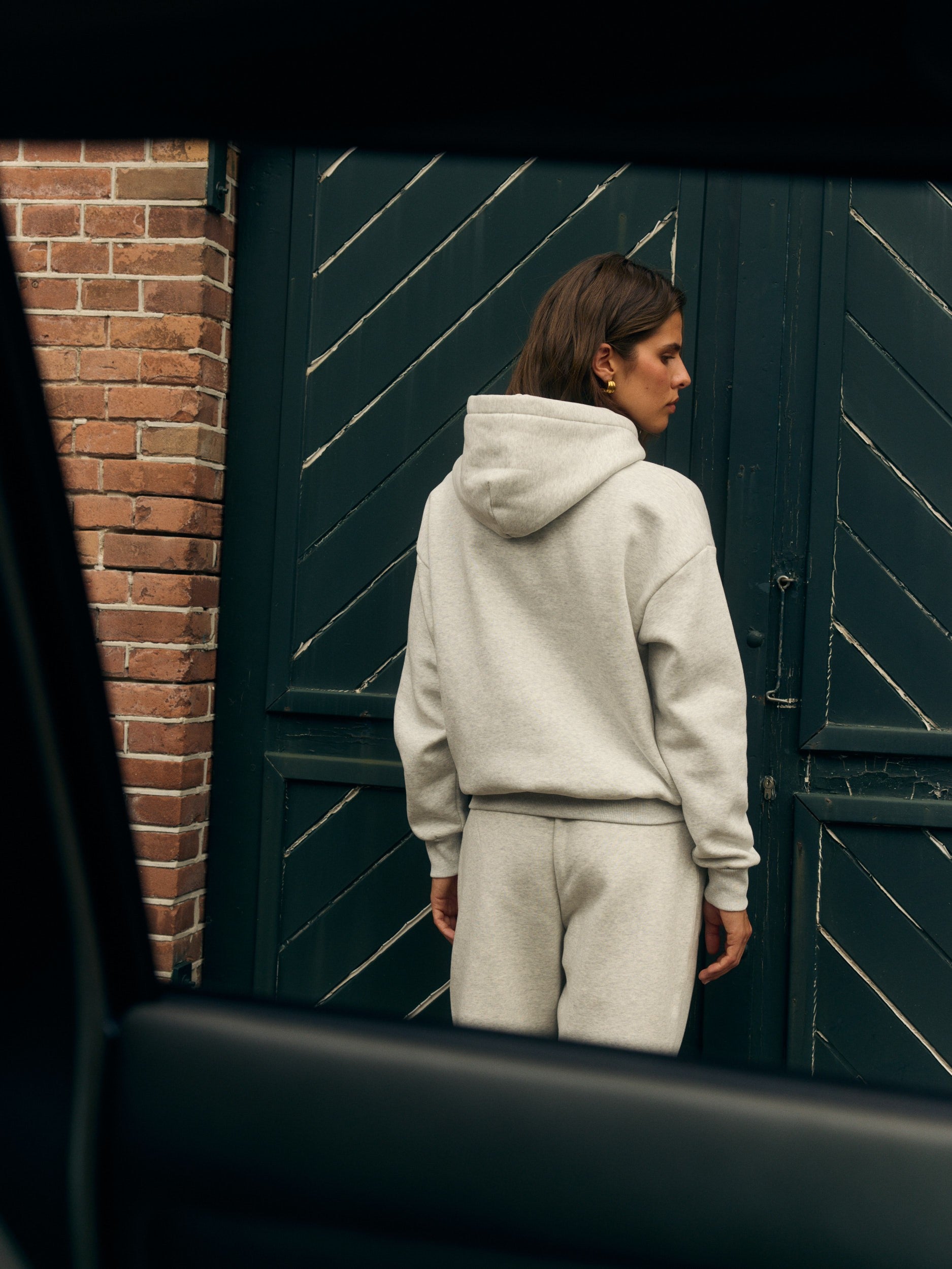Woman wearing a grey melange hoodie and matching sweatpants seen from behind against a dark green door.